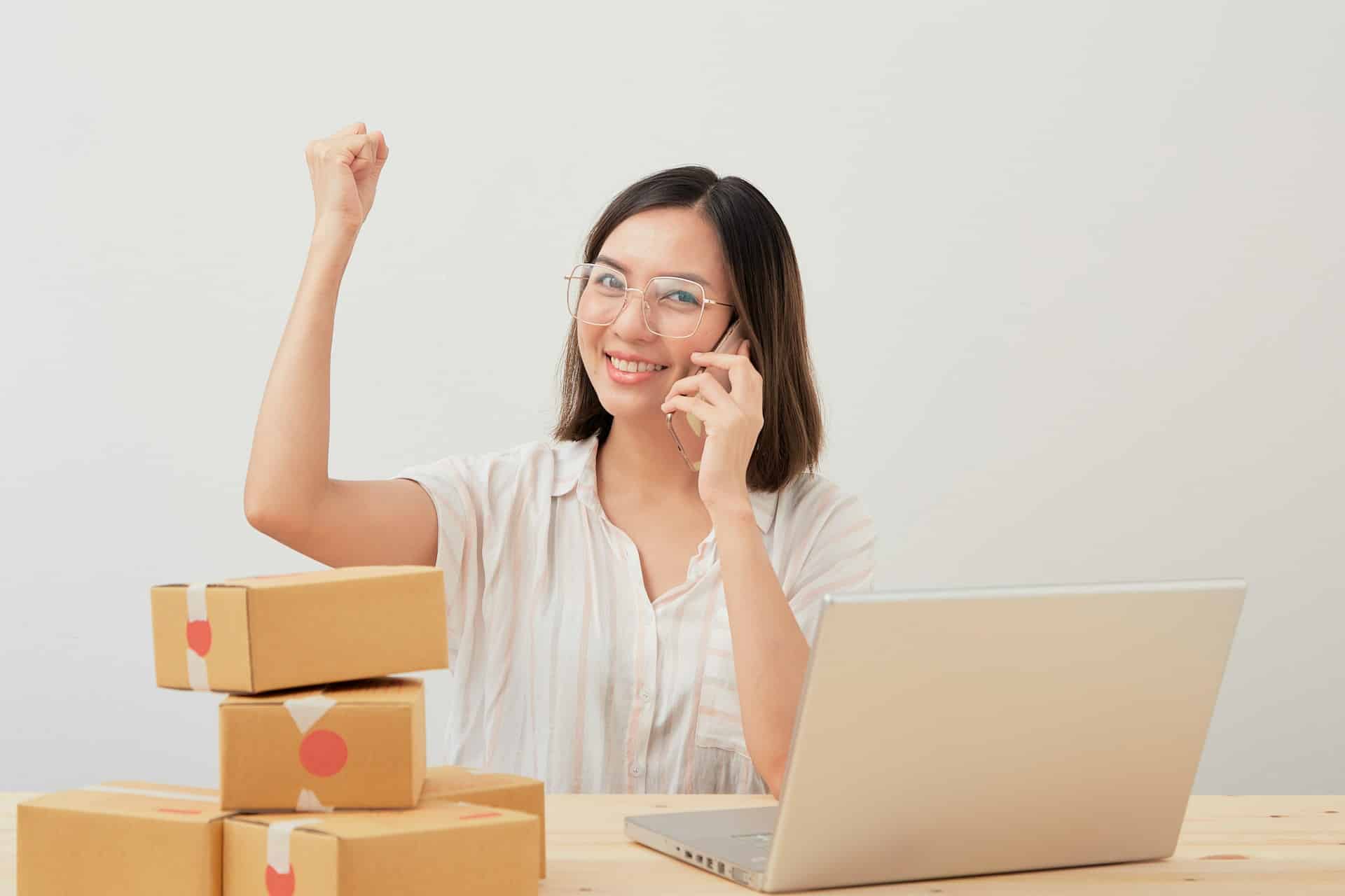 Eine Frau sitz vor einem Laptop, telefoniert und reckt einen Arm vor Freude in die Luft. Neben ihr stehen Pakete